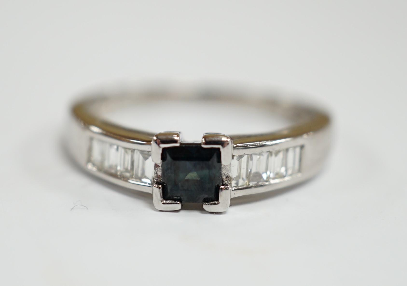 A modern 18ct white gold and single stone square cut sapphire set ring, with baguette cut diamond set shoulders, size N, gross weight 2.9 grams.